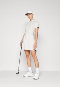 Světlé šedé golfové šaty s límečkem polo, krátkými rukávy a rozevlátou sukní. Obléknuto s bílými teniskami a bílou čepicí, drží golfovou hůl.