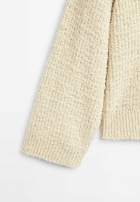 Manche de pull tricoté couleur crème avec un motif gaufré texturé et un poignet côtelé sur un fond blanc.