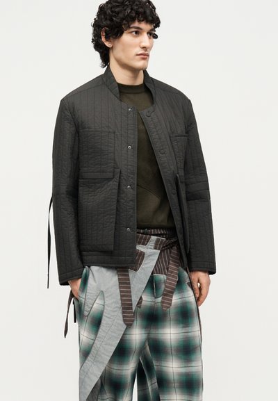 Νέος άνδρας με σγουρά μαλλιά φοράει μαύρο μπουφάν με ραφές πάνω από ένα πράσινο πουλόβερ και φαρδιά καρό παντελόνια με υφασμάτινες λωρίδες σε απλό φόντο.