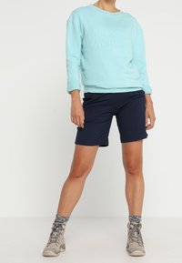 Světle modrá mikina s vyraženým logem, kombinovaná s námořnickými šortkami a béžovými turistickými botami, se šedými vzorovanými ponožkami.