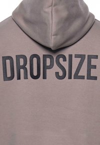 Mikina s kapucí v tlumené růžové látce s výrazným černým textem "DROPSIZE" na zadní straně, s hladkou strukturou a přesným designem.