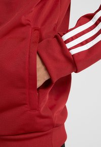 Sudadera roja con franjas blancas en las mangas, tejido texturizado y un bolsillo lateral con abertura oculta.