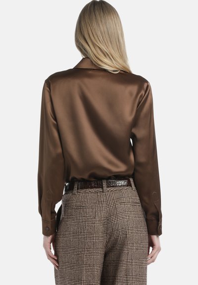 Bluză lungă din satin maro, cu un textură fină, având un guler clasic. Asortată cu pantaloni în carouri cu talie înaltă și o curea lucioasă.