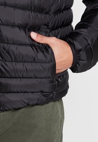 Sort pufferjakke med horisontalt quiltet design. Har en sidelomme, elastiske manchetter og et glat, blankt materiale.