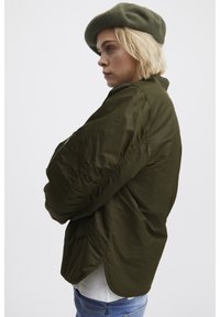 Veste vert olive à coupe décontractée, manches longues, fentes sur les côtés et poche zippée ; portée avec un béret vert assorti.
