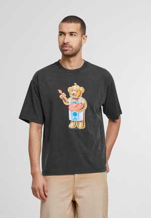 Sorte bomulds T-shirt med et farverigt grafisk tryk af en bjørn, der holder en pensel og en palet, med hvide og blå detaljer på bjørnens tøj.
