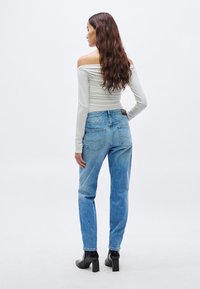 Top gris claro de manga larga y hombros descubiertos combinado con vaqueros azules de talle alto y botas negras de tacón hasta el tobillo. Modelo vista desde atrás.