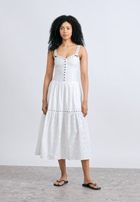 Λευκό, μακρύ φόρεμα χωρίς μανίκια με εφαρμοστό μπούστο, φούστα σε στρώσεις, υφή με τρυπητά σχέδια, μαύρα κουμπιά ως διακοσμητικά και ρυθμιζόμενους ιμάντες στους ώμους.