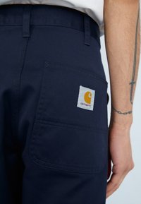 Tamno plave hlače izrađene od izdržljivog materijala, s stražnjim džepom koji ima četvrtasti bijeli tekstilni oznaku i narančastim logotipom. Glatka tekstura.