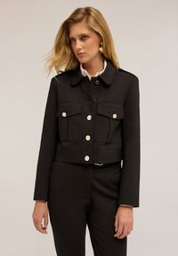 Sort cropped jakke lavet af kraftigt stof, med dobbelt brystlommer, store hvide knapper og krave. Bæres over en hvid skjorte.
