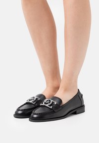 Mocassins en cuir noir avec des accents en métal argenté, semelle plate et détails cousus. Design minimaliste avec bout arrondi et finition lisse.