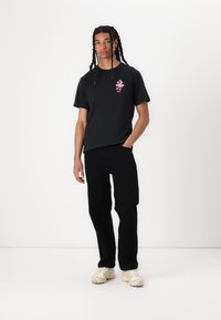 Sort bomulds T-shirt med lyserød grafik på brystet, parret med sorte bukser og lysfarvede sneakers; modellen står med den ene hånd i lommen.