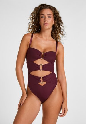 Donna con capelli ricci che indossa un costume intero bordeaux con ritagli e dettagli ad anello dorati su uno sfondo chiaro e neutro.