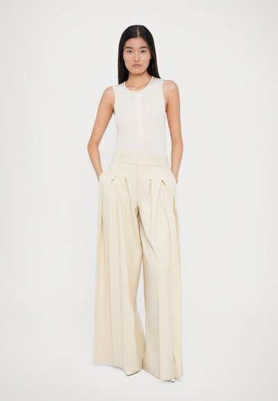 Valkoinen ribattu tanktoppi nappidetailillä, yhdistettynä leveälahkeisiin pleatattuihin housuihin, jotka ovat saman sävyisiä vaalean kermaisia. Sileät tekstuurit.
