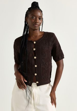 Kvinde med langt flettet hår iført en kortærmet mørkebrun strikket cardigan med fem cremefarvede knapper og hvide bukser.