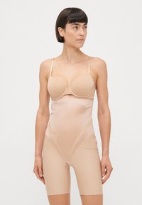 Nøgen shapewear bodysuit med et sømløst design, justerbare stropper og mid-thigh længde. Lavet af glat, elastisk stof.