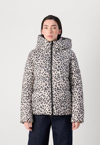 Person in einer leopardenbedruckten Puffjacke mit Kapuze, die vor einem einfarbigen Hintergrund steht, mit sichtbaren Händen an den Seiten.