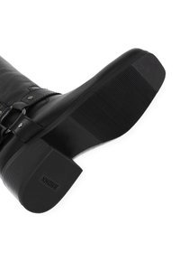 Botte en cuir noir avec une semelle texturée, dotée d'un large talon et d'un détail de boucle réglable. Matériau lisse avec une finition brillante.