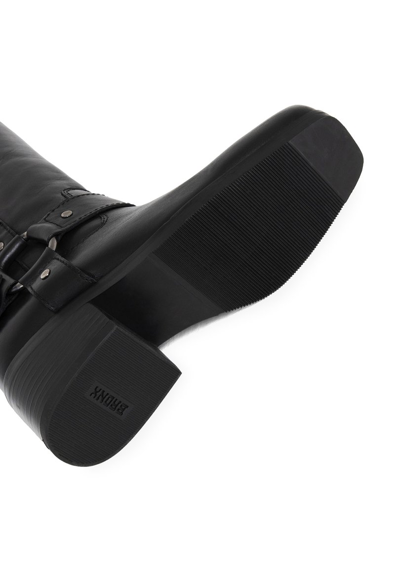 Botte en cuir noir avec une semelle texturée, dotée d'un large talon et d'un détail de boucle réglable. Matériau lisse avec une finition brillante.