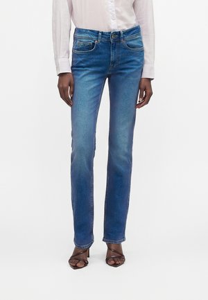 Femme portant un jean bleu droit taille mi-haute, une chemise blanche à manches longues et des sandales à talons ouvertes marron, debout sur fond blanc.