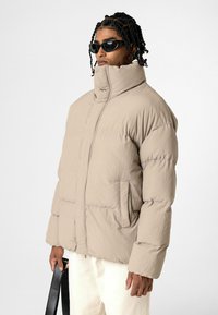 Beige pufferjack met hoge kraag, gestikte textuur, rits aan de voorkant en zijzakken. Model draagt zwarte zonnebril en houdt een tas vast.