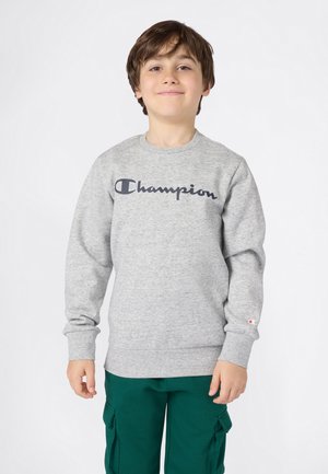 Ragazzo che indossa una felpa grigia Champion e pantaloni cargo verdi, in piedi e sorridente contro uno sfondo bianco puro.