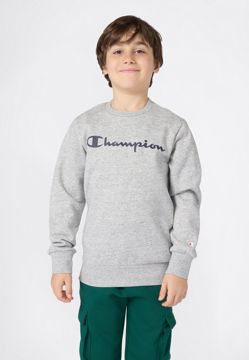 Ragazzo che indossa una felpa grigia Champion e pantaloni cargo verdi, in piedi e sorridente contro uno sfondo bianco puro.