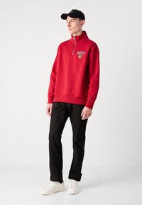 Hanorac roșu cu fermoar, cu logo brodat, tiv și manșete ribbate, purtat cu pantaloni negri și adidași albi. Modelul poartă o șapcă neagră.
