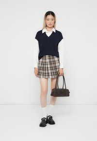 Gilet tricoté bleu marine sur une chemise blanche à col, jupe à carreaux dans des tons bruns, sac à main sombre, chaussettes blanches, chaussures à brides noires. Design simple.