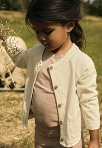 Cardigan tricoté blanc avec des boutons en bois, associé à un haut rose clair avec un motif texturé. Herbe verte et foin en arrière-plan.