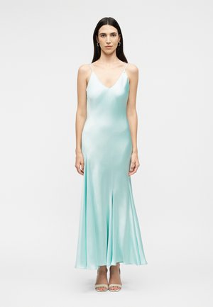 Γυναίκα που φοράει ανοιχτό γαλάζιο σατέν φόρεμα με λεπτές τιράντες, όρθια σε απλό λευκό φόντο.