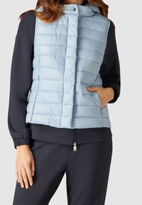 Ljusblå vadderad väst med horisontellt quiltat mönster, framknäppning och sidofickor, bärs över en mörk sweatshirt.