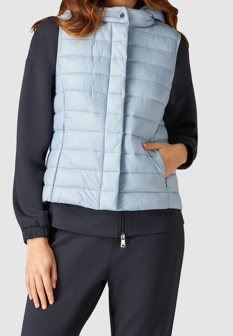 Ljusblå vadderad väst med horisontellt quiltat mönster, framknäppning och sidofickor, bärs över en mörk sweatshirt.