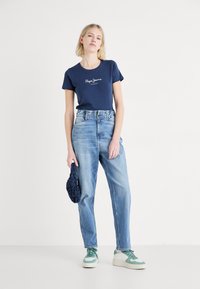 Navyblauw t-shirt met wit logo, lichtblauwe hoge taille jeans, groen en witte sneakers, met een gestructureerde donkerv blauw tas.