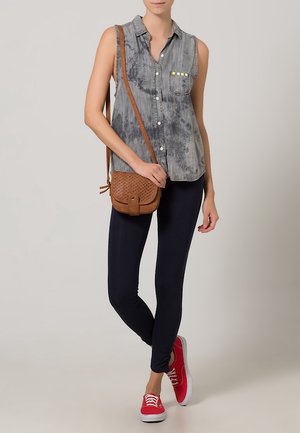 Woman wearing sleeveless gray button-up shirt, black leggings, red sneakers, and carrying a brown crossbody bag against a plain background.