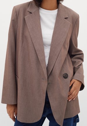 Persoon in een losse bruin blazer over een wit t-shirt en blauwe jeans, met één hand op de blazerzak.