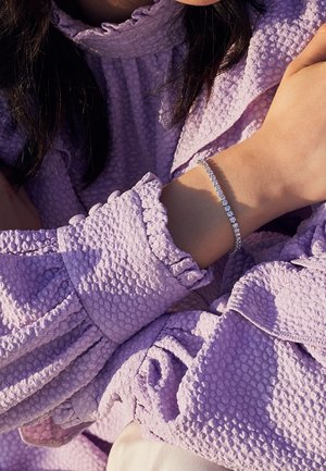 Vrouw draagt een gestructureerde lavendelkleurige blouse met gerimpelde manchetten en een tennisarmband om haar pols in natuurlijk licht.