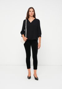 Black, loose-fitting V-neck blouse with three-quarter sleeves paired with black, fitted pants. Model holds a small white handbag.