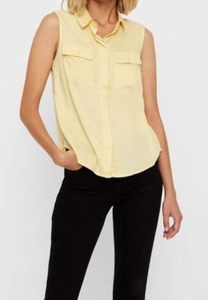 Bezrekawowa, jasnożółta bluzka z kołnierzykiem i przednimi kieszeniami, noszona z czarnymi spodniami, modelka pozująca z jedną ręką w kieszeni.