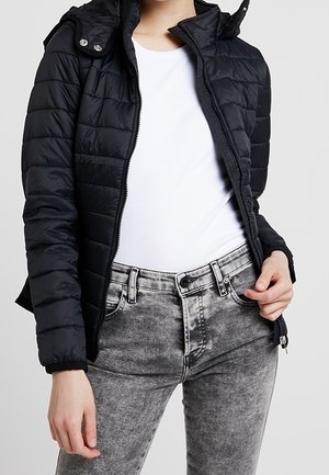 Kobieta ubrana w czarną pikowaną kurtkę na białej koszulce i szare jeansy z efektem acid wash, trzyma jedną ręką brzeg kurtki.