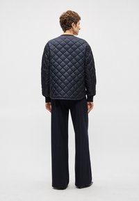 Jūras zila quiltota jaka ar garām piedurknēm un ribotām aprocēm; apvienota ar jūras zila svītrainām platiem biksēm. Tekstūrēts audums ar dimanta modeli.