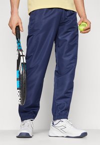 Marineblå joggebukser med elastisk midjebånd, sammen med hvite joggesko med marineblå detaljer. Personen holder en tennisracket og en ball.