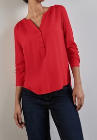Rote Bluse mit langen Ärmeln und V-Ausschnitt, Knopfdetails und abgerundetem Saum. Der Stoff wirkt glatt mit einer leicht glänzenden Textur. Getragen mit dunklen Jeans.