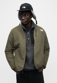 Άνδρας με πράσινο μπουφάν με φερμουάρ, γκρι φλις πουλόβερ και μαύρο χειμερινό καπέλο The North Face με καλύμματα αυτιών, τα χέρια στις τσέπες, κοιτάζοντας προς τα δεξιά.