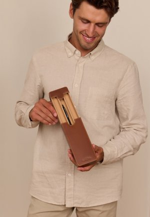 Muž v béžovej košeli drží otvorené hnedé kožené púzdro na pero s čiernym perom vo vnútri, usmieva sa a pozerá sa dole na púzdro.