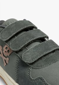 Zapatilla de ante gris con dos correas de Velcro; presenta un diseño de calavera y huesos cruzados en marrón. Suela de goma lisa y detalles cosidos.