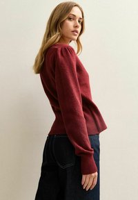 Burgundowy sweter z bufiastymi rękawami i prążkowanymi mankietami, zestawiony z ciemnymi dżinsami. Miękka tekstura z lekko rozkloszowanym dołem.