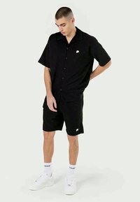Maglietta nera a maniche corte con bottoni, abbinata a pantaloni corti neri. Presenta un piccolo logo sul lato sinistro del petto e sneakers bianche.