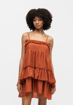 Νεαρή γυναίκα με σγουρά μαλλιά που φορά ένα αμάνικο, στρωμένο φόρεμα σε σκουροπορτοκαλί απόχρωση με λεπτομέρειες βολάν, μπροστά σε μονόχρωμο φόντο.