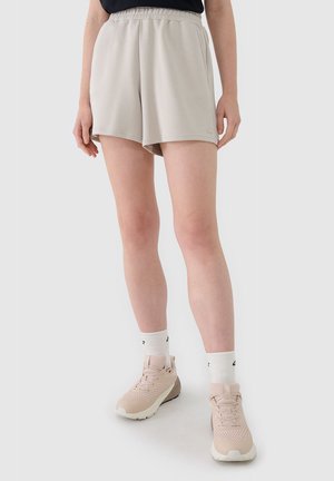 Mujer con pantalones cortos beige de cintura elástica, calcetines blancos y tenis deportivos rosa claro, de pie contra un fondo claro y liso.